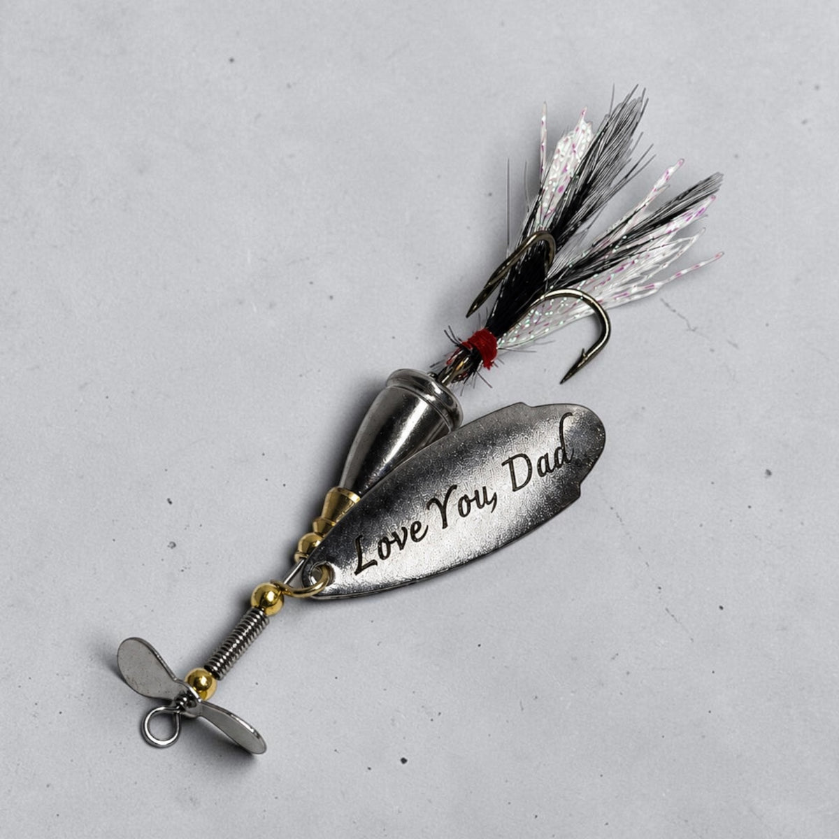 A custom metal fishing lure engraved with 'Love You, Dad' on its main blade, featuring a silver spinner blade, a split blade, gold-colored beads, a black and white feathered jig head, and a red accent, photographed on a neutral gray background.