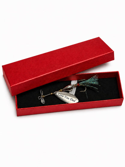 A silver metal fishing lure engraved with 'Love You Dad' rests inside an open red presentation box with a black velvet lining, with the matching red lid propped behind it on a white background.