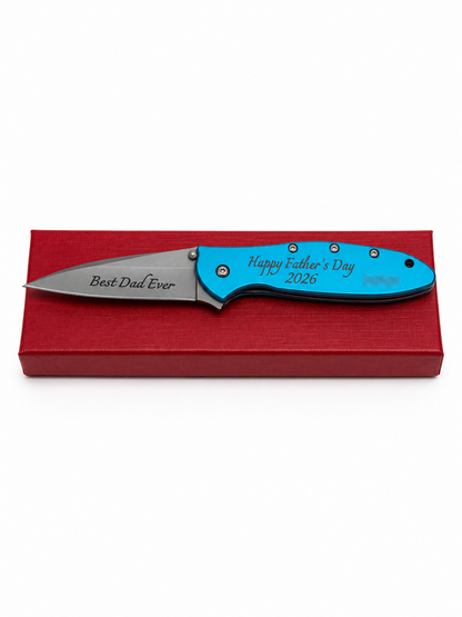 A personalized folding pocket knife with a bright blue handle and a silver blade, resting on a red gift box. The handle is engraved with 'Happy Father's Day 2026' and the blade is engraved with 'Best Dad Ever'.