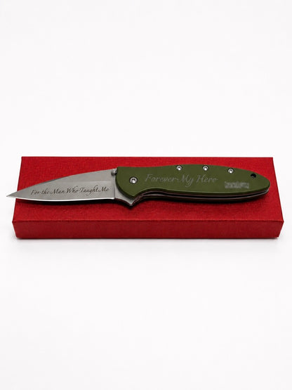An engraved folding pocket knife with an olive green handle and a silver stainless steel blade, resting on a red presentation box against a white background.