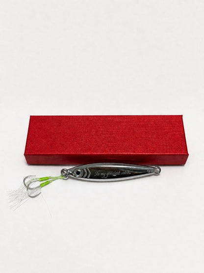 A silver metal fishing lure with green and white skirt strands, displayed horizontally in front of a textured red rectangular box on a plain white background.