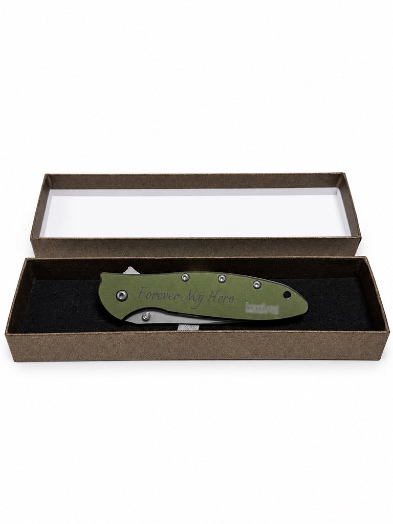 An olive green folding pocket knife with 'Compton, CA, 9000' engraved on the handle, presented inside an open brown gift box with a clear lid on a white background.