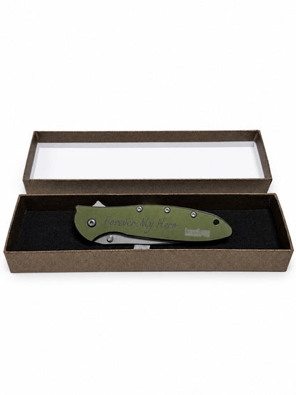 An olive green folding pocket knife with 'Compton, CA, 9000' engraved on the handle, presented inside an open brown gift box with a clear lid on a white background.