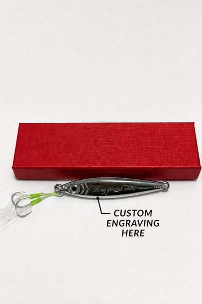 A silver metal fishing lure with green and white fishing line and hooks, shown in front of a textured red box on a white background. A black text overlay reads 'CUSTOM ENGRAVING HERE' with an arrow pointing to the lure's body.