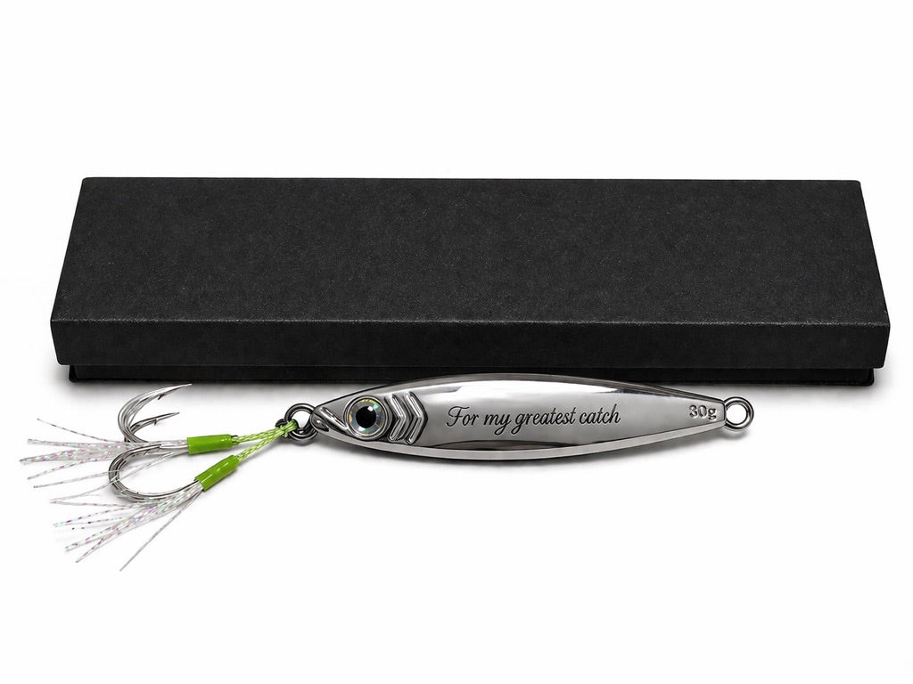 A silver metal fishing lure engraved with 'For my greatest catch' and a green-and-silver skirt, displayed in front of a black presentation box on a white background.