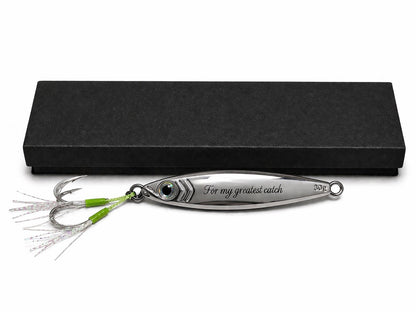 A silver metal fishing lure engraved with 'For my greatest catch' and a green-and-silver skirt, displayed in front of a black presentation box on a white background.