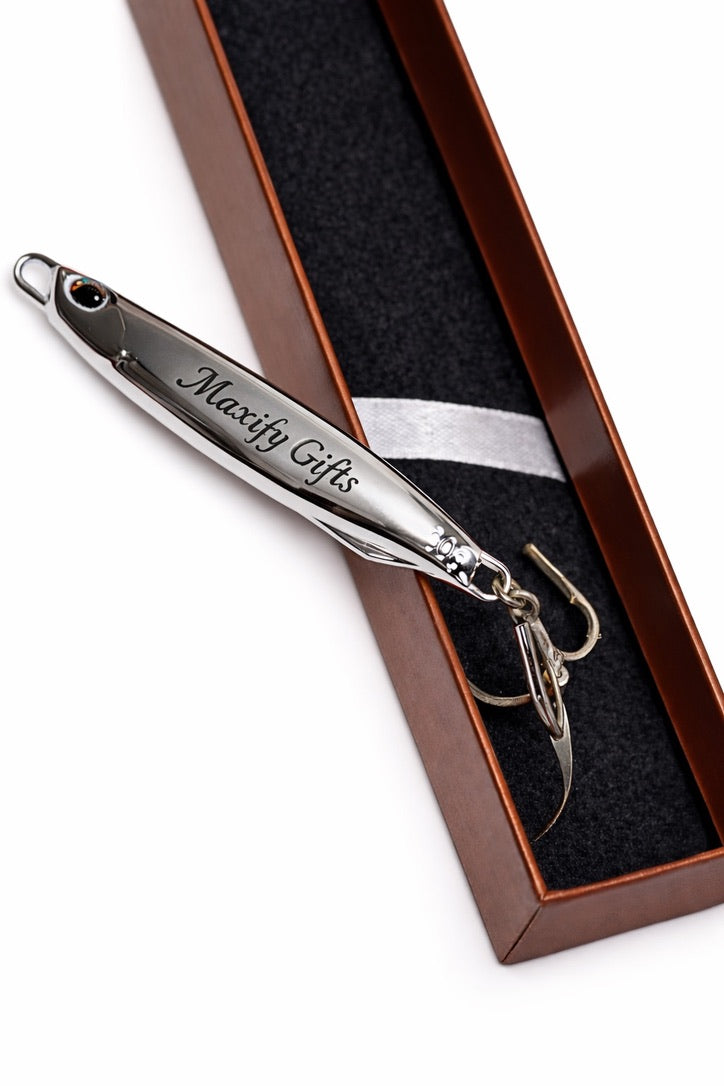 A silver metal novelty fishing lure engraved with 'Maxify Gifts' and a treble hook, presented inside a brown gift box with a black velvet insert on a white background.