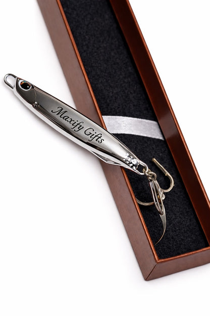 A silver metal novelty fishing lure engraved with 'Maxify Gifts' and a treble hook, presented inside a brown gift box with a black velvet insert on a white background.