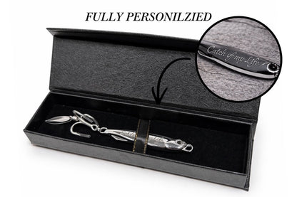 A silver metal fishing lure presented inside an open black gift box with a black velvet interior. A circular inset shows a close-up of the lure handle with engraved text reading 'Catch of my life!'.