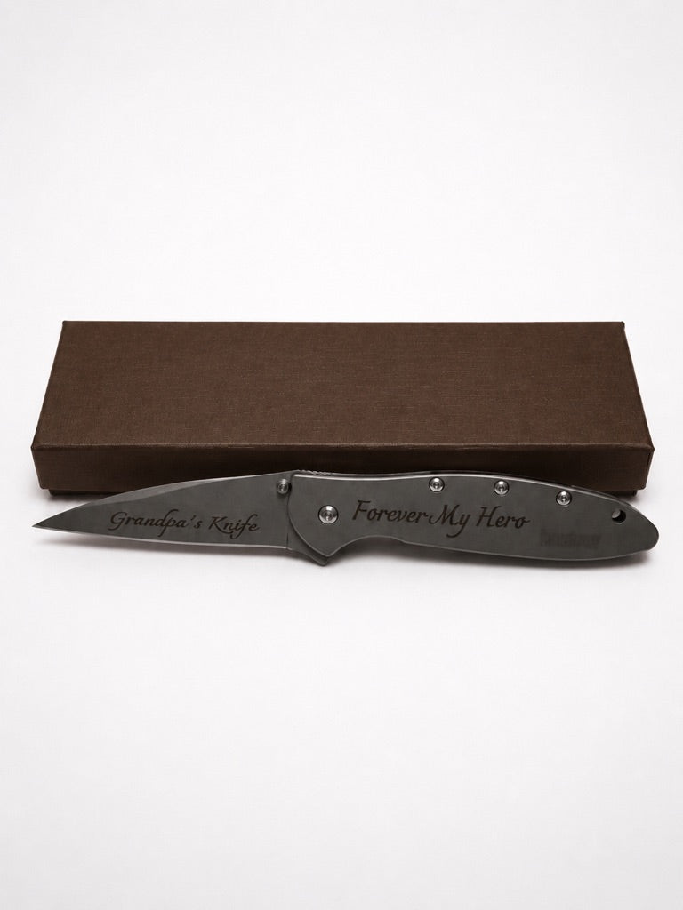 A personalized folding pocket knife with a dark gray metal blade and handle, displayed in front of its brown presentation box on a white background. The blade and handle are engraved with 'Grandpa's Knife' and 'Forever My Hero'.