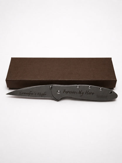 A personalized folding pocket knife with a dark gray metal blade and handle, displayed in front of its brown presentation box on a white background. The blade and handle are engraved with 'Grandpa's Knife' and 'Forever My Hero'.