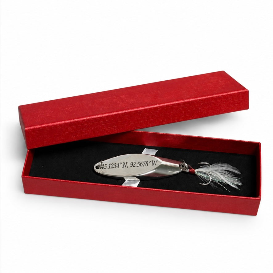 A silver metal fishing lure engraved with geographic coordinates, presented inside an open red gift box with a black interior and a red lid on a white background.