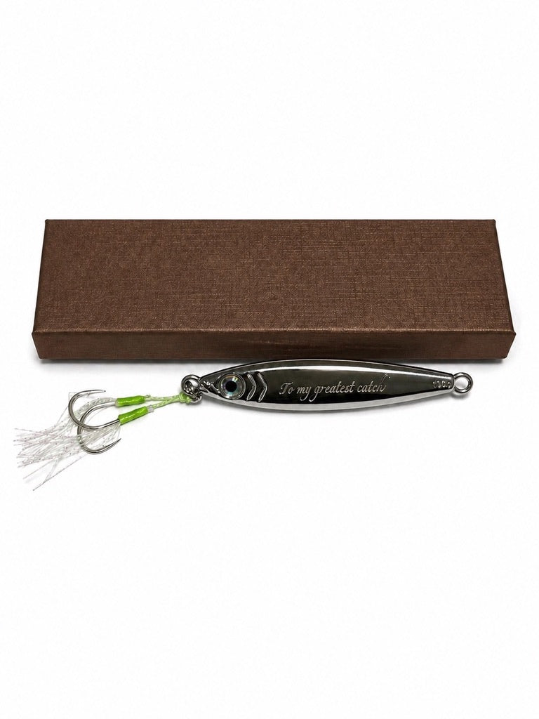 A silver metal fishing lure engraved with 'To my grateful wife' and attached to two green-tipped strands with white tinsel, displayed in front of a brown textured gift box on a white background.