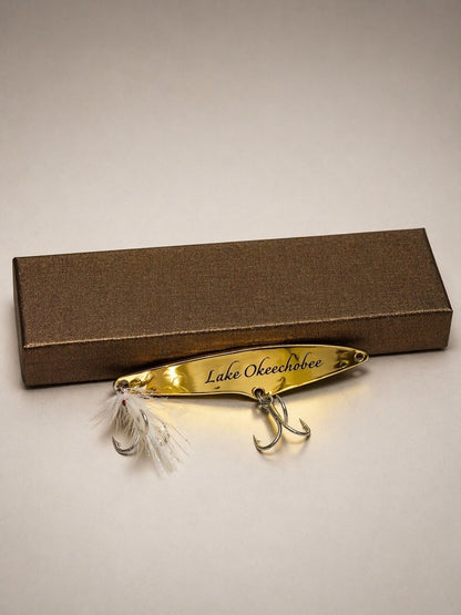 A gold metal fishing lure engraved with 'Lake Okeechobee' and featuring white feathers and silver hooks, displayed in front of a textured brown gift box on a neutral light gray background.