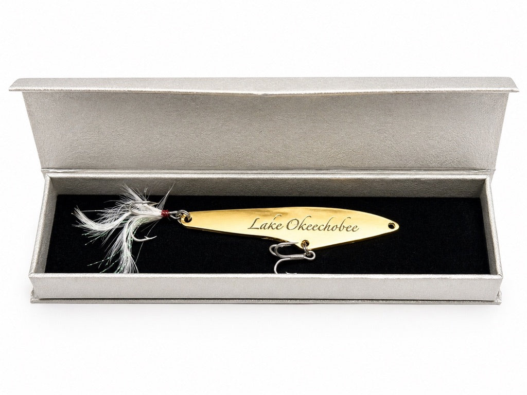 A gold metal fishing lure engraved with 'The Lake Okeechobee' and displayed inside an open silver presentation box with a black velvet insert.
