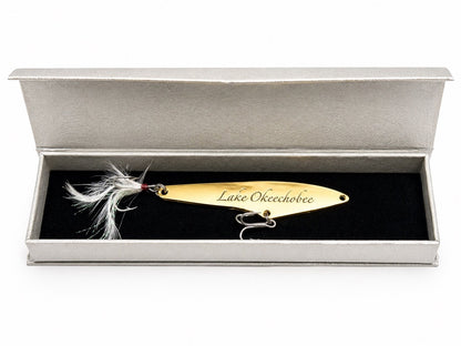 A gold metal fishing lure engraved with 'The Lake Okeechobee' and displayed inside an open silver presentation box with a black velvet insert.