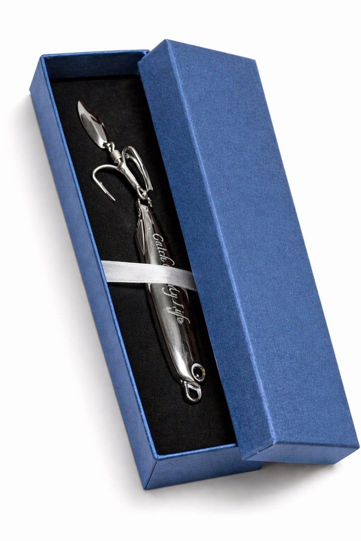 A silver metal fishing lure with hooks attached, presented inside an open blue gift box with a black insert and a white ribbon, on a white background.
