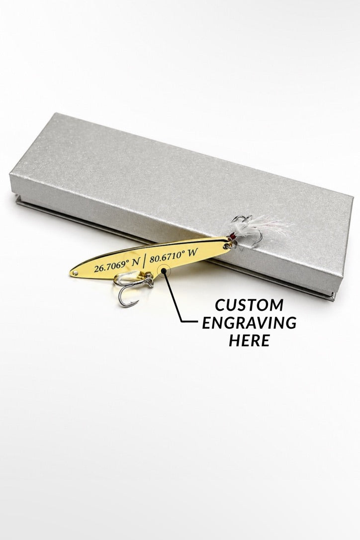 A gold-colored metal fishing lure with custom coordinates engraved on its side, shown next to a textured silver gift box on a white background. A black line points to the engraving with the text 'CUSTOM ENGRAVING HERE'.