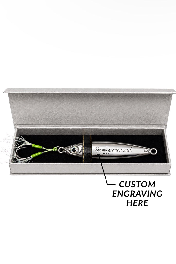 A silver metal fishing lure displayed inside an open gray presentation box with black lining. The lure has a bright green skirt and wire hooks, and the box is labeled 'CUSTOM ENGRAVING HERE' with an arrow pointing to the engraved area on the lure.