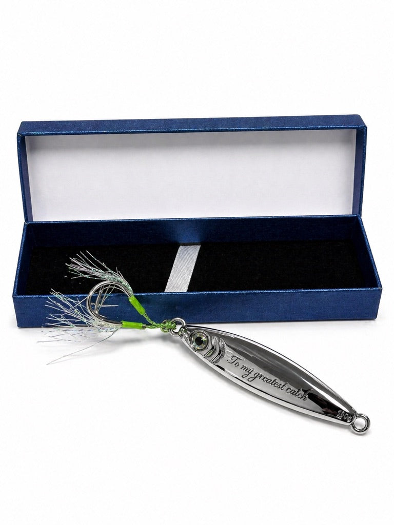 A silver metal fishing lure with a green and white feather skirt, displayed in front of its open dark blue presentation box on a white background.