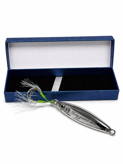 A silver metal fishing lure with a green and white feather skirt, displayed in front of its open dark blue presentation box on a white background.