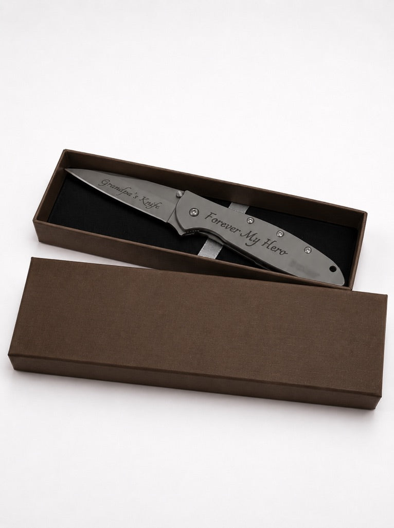 A personalized folding pocket knife with a brushed silver finish and engraved text, presented in an open brown gift box with its lid placed below.