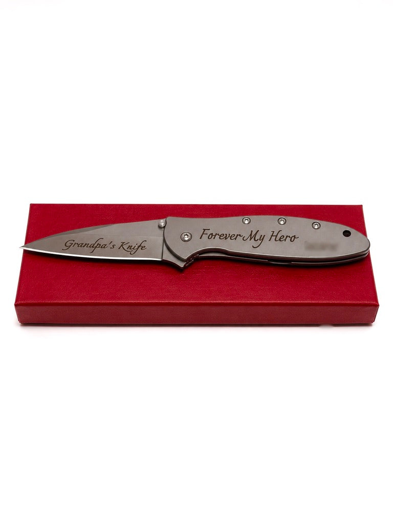 A personalized folding pocket knife with a satin finish, featuring 'Grandpa's Knife' on the blade and 'Forever My Hero' on the handle, resting on a red gift box against a white background.