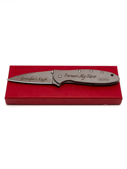 A personalized folding pocket knife with a satin finish, featuring 'Grandpa's Knife' on the blade and 'Forever My Hero' on the handle, resting on a red gift box against a white background.