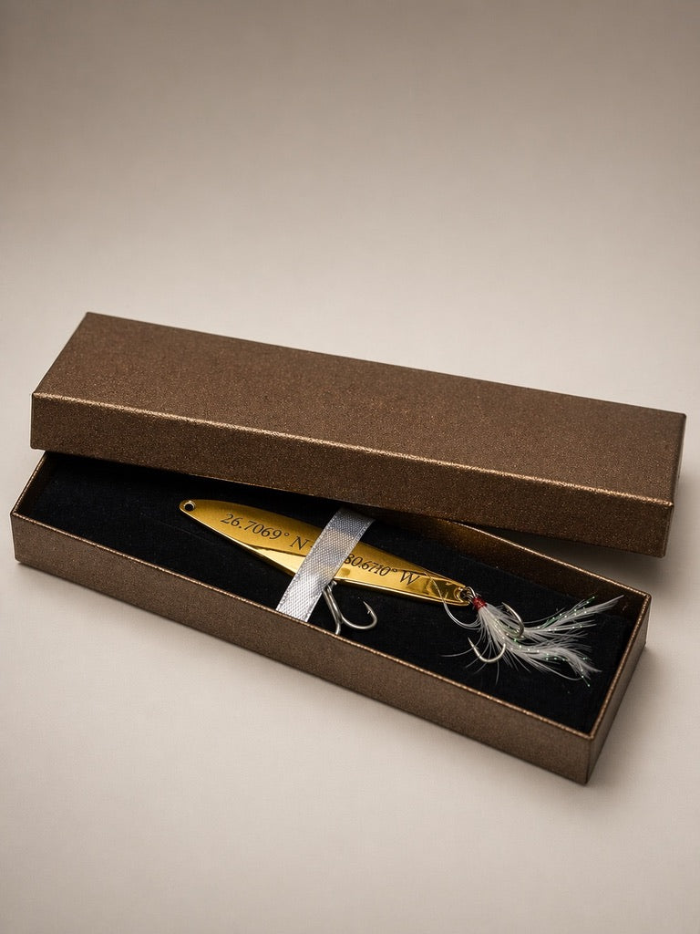A gold metal fishing lure with white and gray feather accents, presented inside an open textured brown gift box with a black velvet interior.