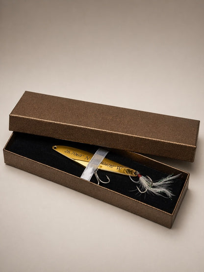 A gold metal fishing lure with white and gray feather accents, presented inside an open textured brown gift box with a black velvet interior.