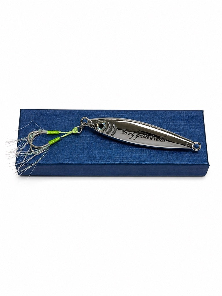 A silver metal fishing lure engraved with 'To my greatest catch' resting on a textured blue presentation box against a white background.