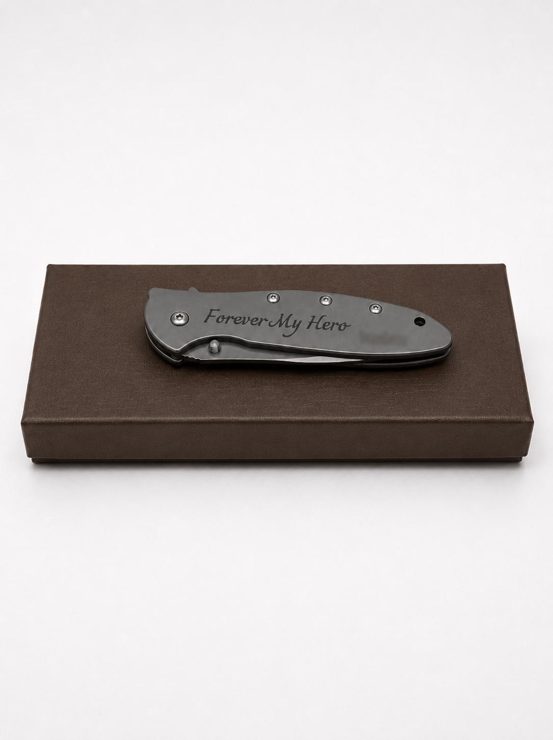 A black folding pocket knife with 'Forever My Hero' engraved on the handle, resting on a brown presentation box against a white background.