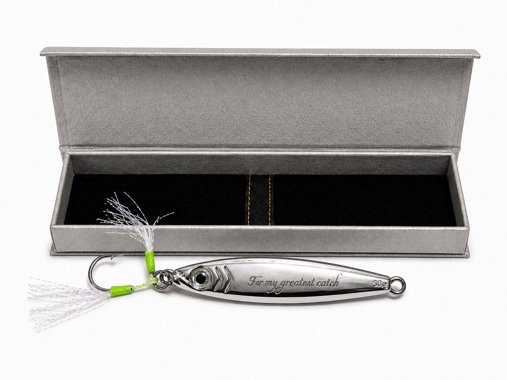 A silver metal fishing lure engraved with 'For my greatest catch' and '300 g', displayed in front of an open textured gray presentation box with a black interior.