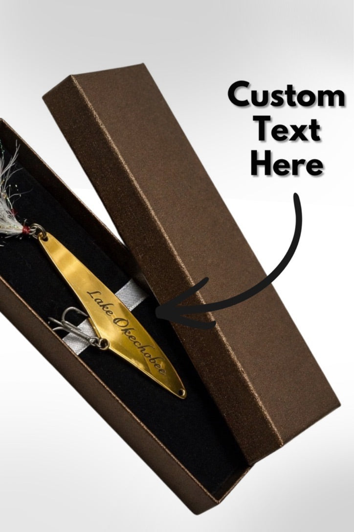 A gold custom-etched fishing lure displayed inside an open brown presentation box with a black insert, shown on a light gray background with a 'Custom Text Here' text overlay and arrow.