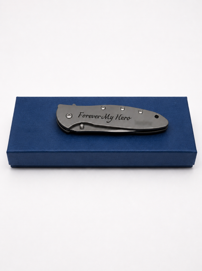 A dark gray folding pocket knife with 'Forever My Hero' engraved on the handle, resting on a dark blue presentation box against a white background.