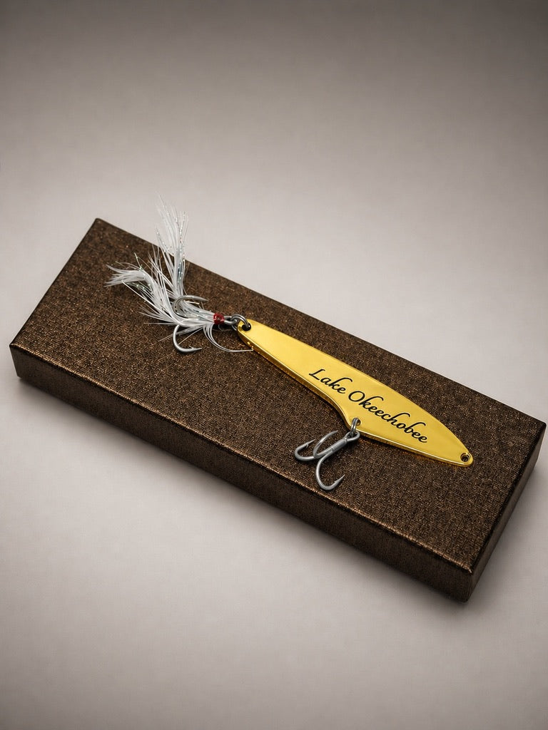 A yellow metal fishing lure with 'Lake Ok pochonk' engraved on the side, displayed on a textured brown presentation box against a neutral gray background.
