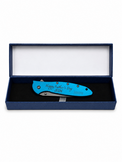 A blue folding pocket knife with 'Happy Father's Day 2026' engraved on the handle, presented inside an open dark blue gift box with a white interior.