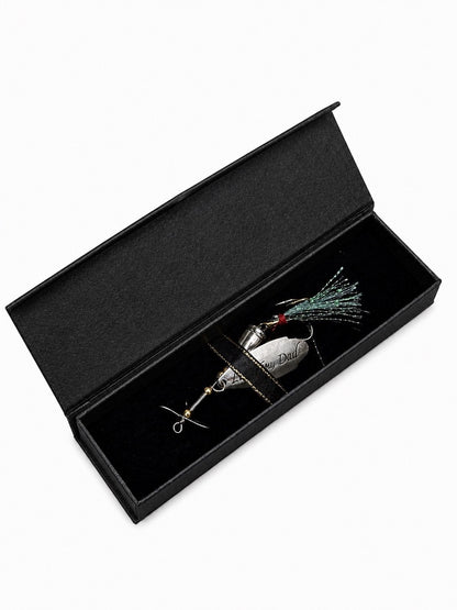 A silver metal fishing lure with a green and black skirt, presented inside an open black velvet gift box on a white background.