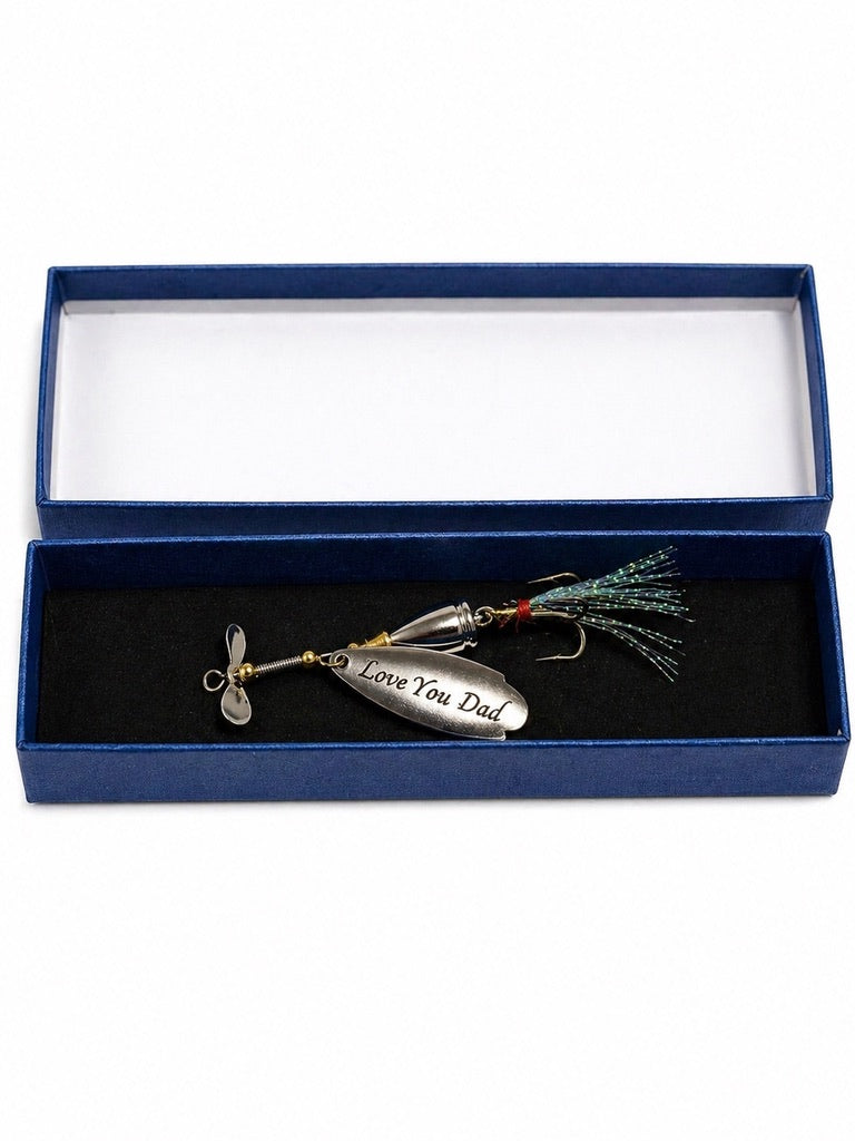 A silver metal fishing lure engraved with 'Love You Dad' and a small propeller, presented inside an open dark blue gift box with a black velvet insert.