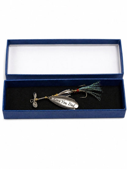A silver metal fishing lure engraved with 'Love You Dad' and a small propeller, presented inside an open dark blue gift box with a black velvet insert.