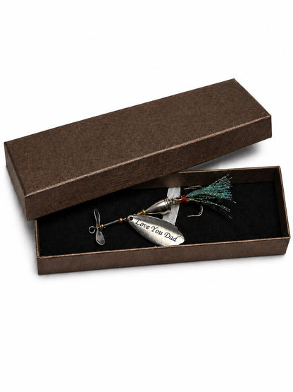 A silver metal fishing lure engraved with 'Love You Dad' rests inside an open brown presentation box with a black velvet lining. The box lid is propped behind it on a clean white background.
