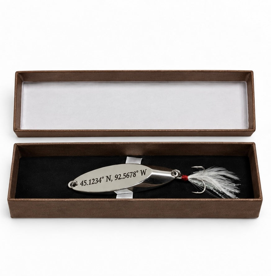 A silver metal fishing lure engraved with geographic coordinates, displayed inside an open brown presentation box with a black velvet insert.