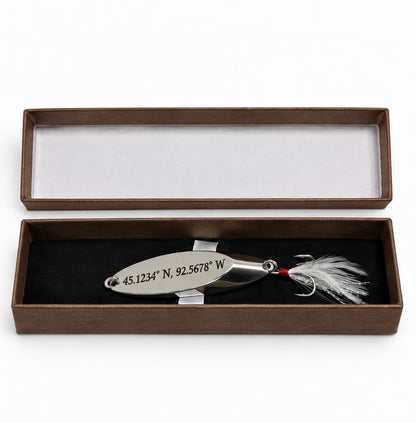 A silver metal fishing lure engraved with geographic coordinates, displayed inside an open brown presentation box with a black velvet insert.