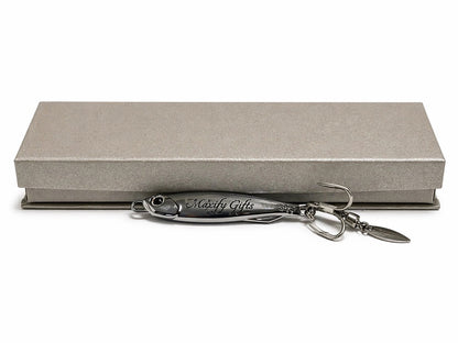 A silver metal fishing lure engraved with 'Moxify Gifts' resting in front of a textured gray presentation box on a white background.