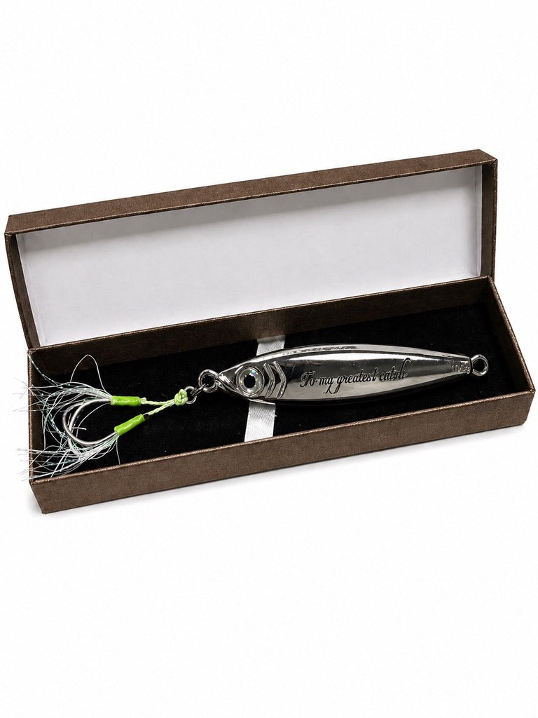 A silver metal fishing lure with a green and white skirt, presented inside an open brown gift box with a white interior.