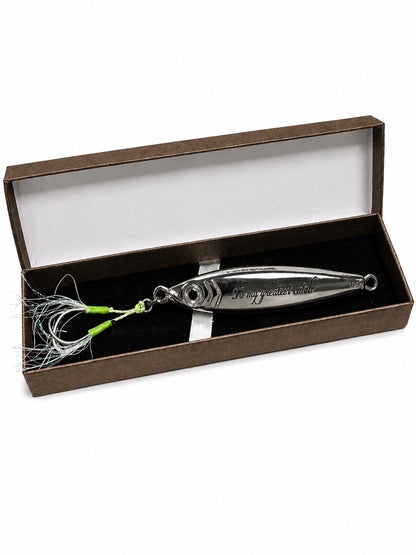 A silver metal fishing lure with a green and white skirt, presented inside an open brown gift box with a white interior.