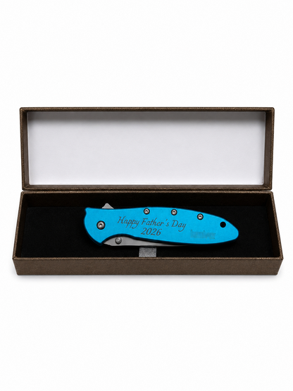 A bright blue folding pocket knife with 'Happy Father's Day 2026' engraved on the handle, presented inside an open brown gift box with a black velvet insert.