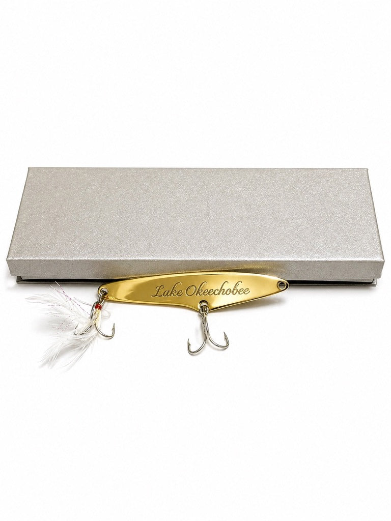 A gold metal fishing lure engraved with 'Lake Opechota' and featuring two treble hooks and a white feather fly, displayed in front of a textured silver gift box on a white background.