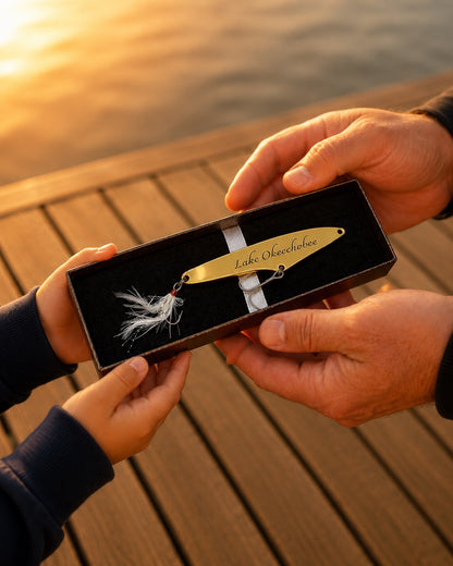 A person holding an open presentation box containing a gold fishing lure with white feather detailing and a silver ribbon. The box is engraved with the text 'Lake Outdoors'.