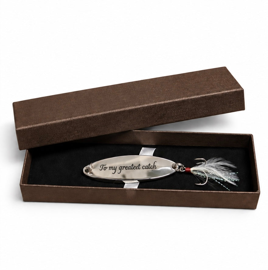 A silver metal fishing lure engraved with 'To my greatest catch' in black script, displayed inside an open brown presentation box with a black insert. The lure is equipped with white and silver feather strands and a small red bead near the hook.